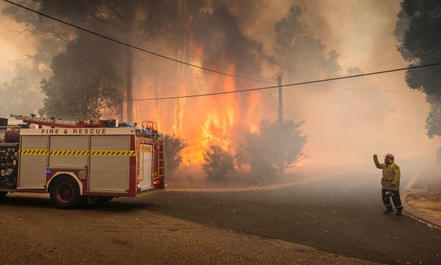A firefighter and fire tanker dwarfed by a raging bushfire. 