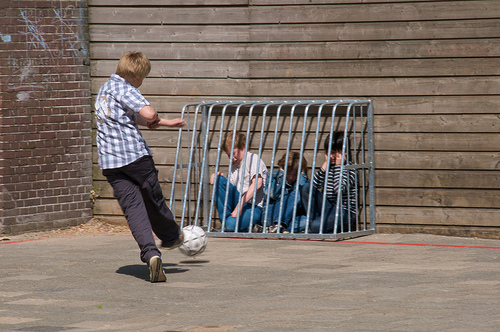 Large buy kicks socccer ball at younger boys cowering in a cage.